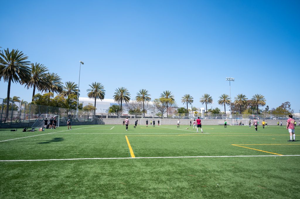 Photo of a soccer field surrounded by palm trees and airport parking lots. Players in purple Hawaiian shirts and black shirts are playing on the field.