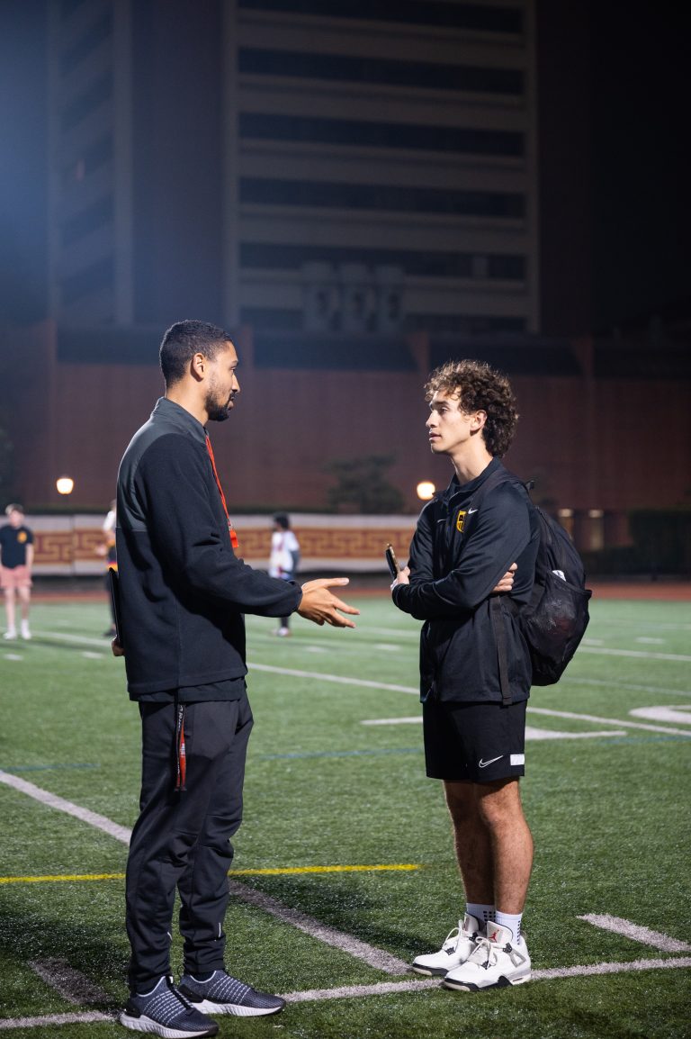 Two men talk to each other on a soccer field. One has a backpack on.