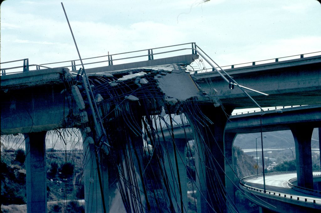 A collapsed freeway bridge with a large amount of damaged rebar hanging off the edge.